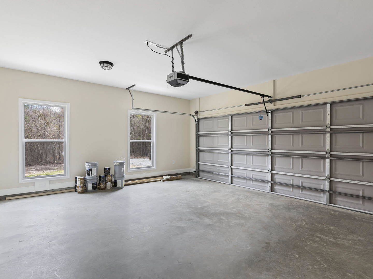 Spacious garage with plaster walls, concrete floor, high ceiling, large window overlooking trees, garage door with attached pipe, and grey cylinder with white label