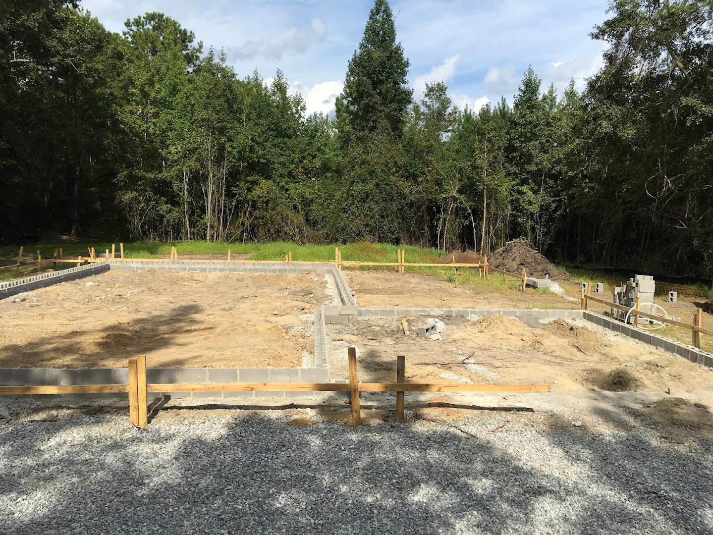Gravel construction site bordered by a wooden fence, piles of gravel and lumber, group of trees in the background under blue sky with clouds, shadow of a dog cast on the ground