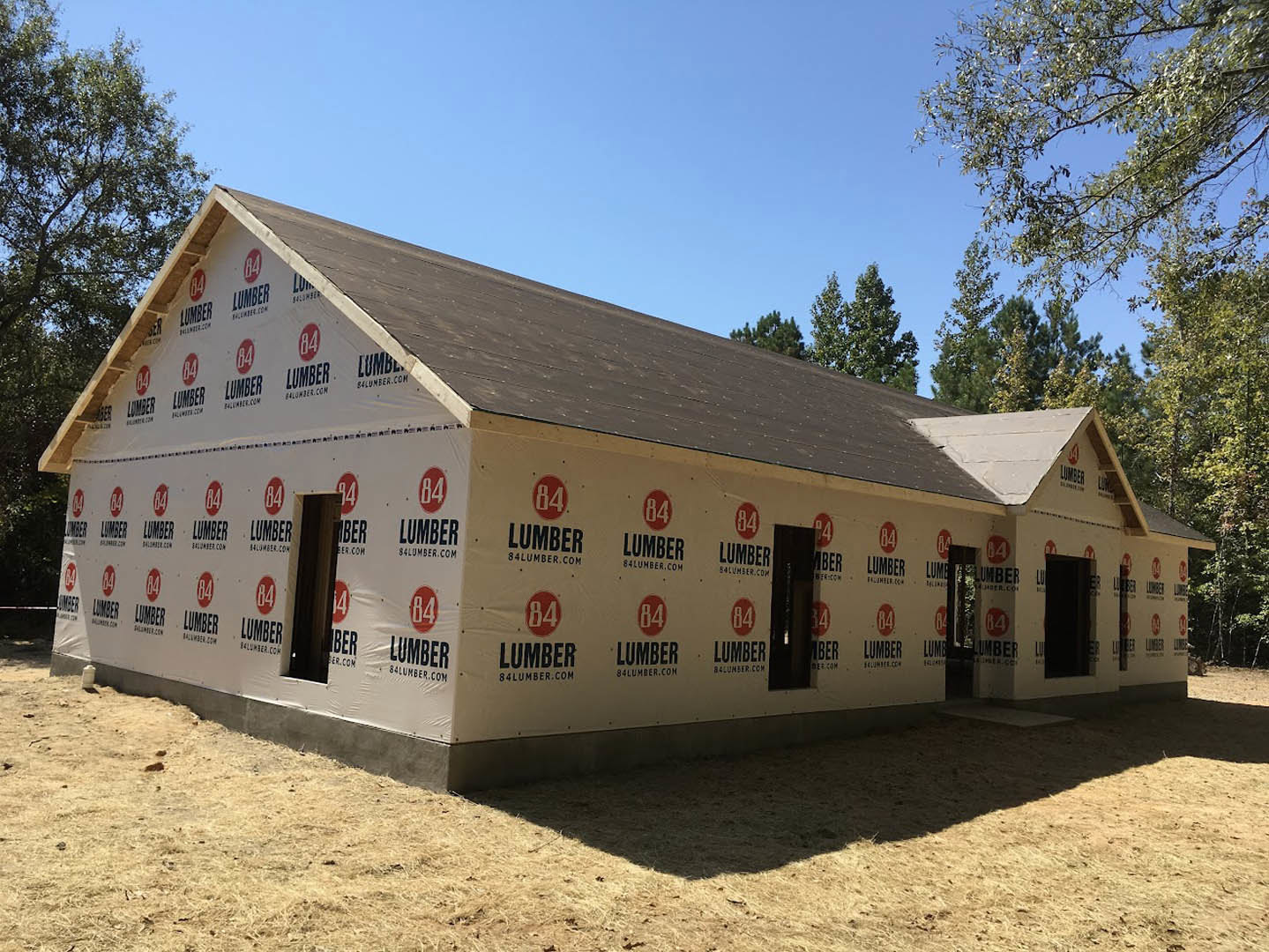 Framed house under construction with exposed wooden beams, surrounded by trees and blue sky in the background