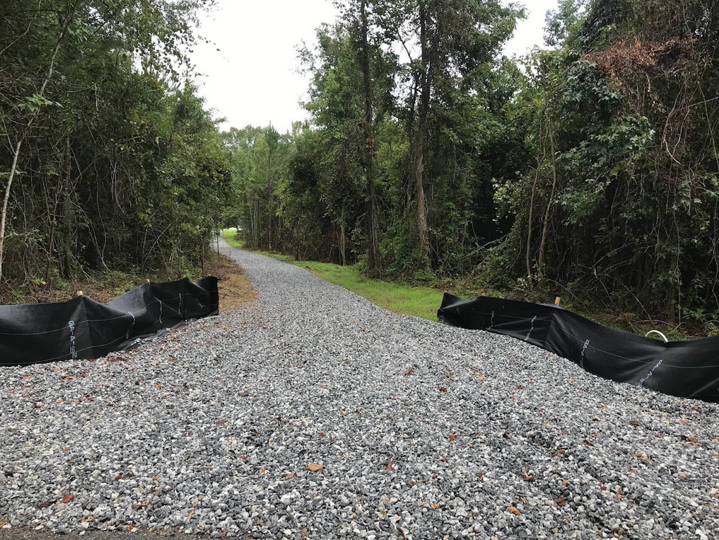 Gravel driveway bordered by grass and trees, black tarps laid over sections of the road, forested area in the background