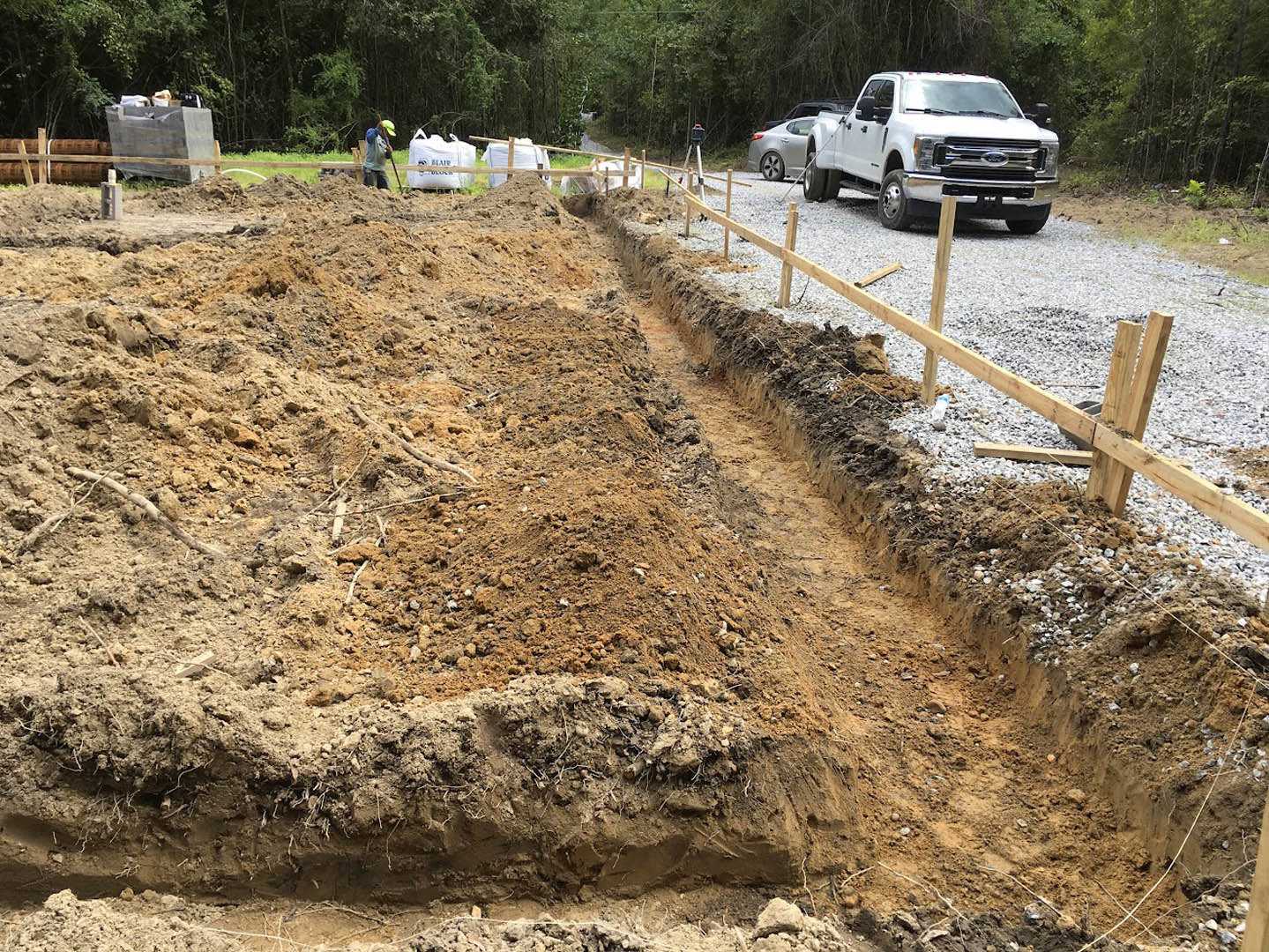 White pickup truck parked on gravel road beside wooden fence, dirt trench with two men standing nearby, large gray box and pile of trash, trees in background.