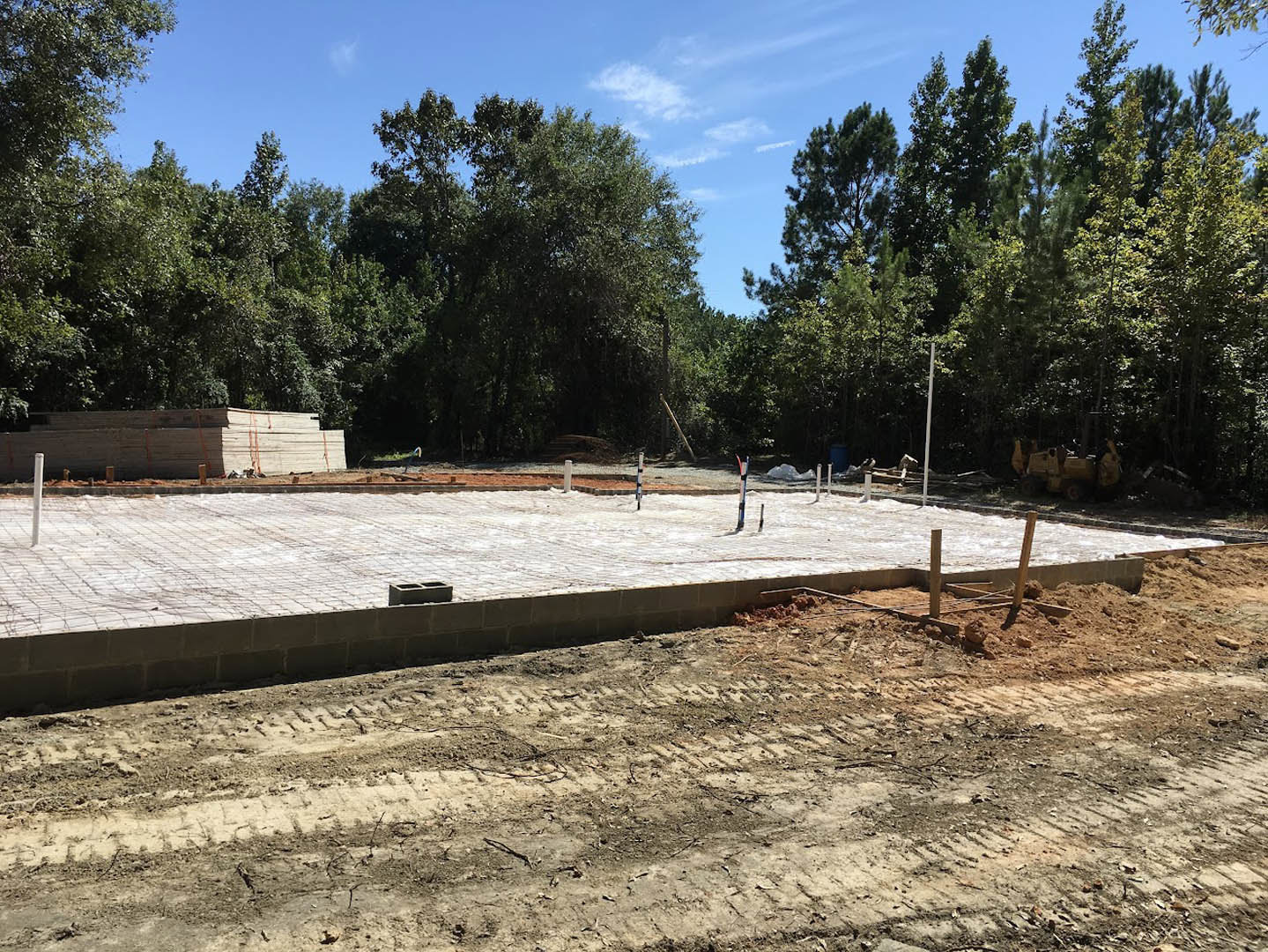 Dirt construction site bordered by leafy trees under a blue sky, with a wooden beam, brick patio, and utility pole visible