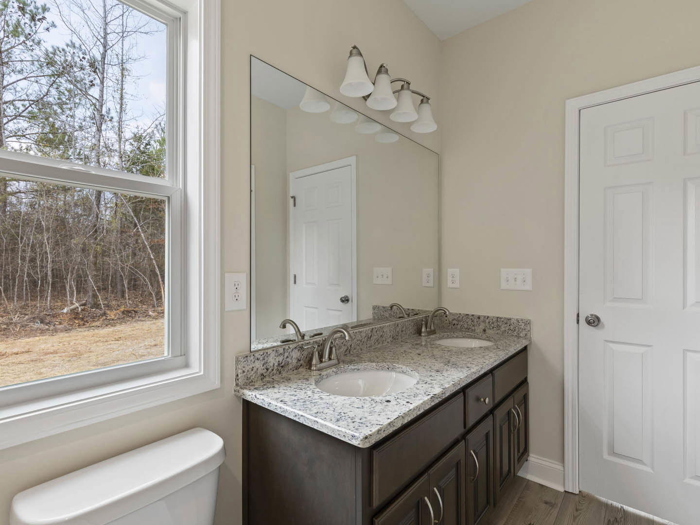 Modern bathroom featuring a rectangular mirror above a white sink with chrome faucet, white toilet, tiled floor, white cabinetry, window overlooking trees, white door with metal