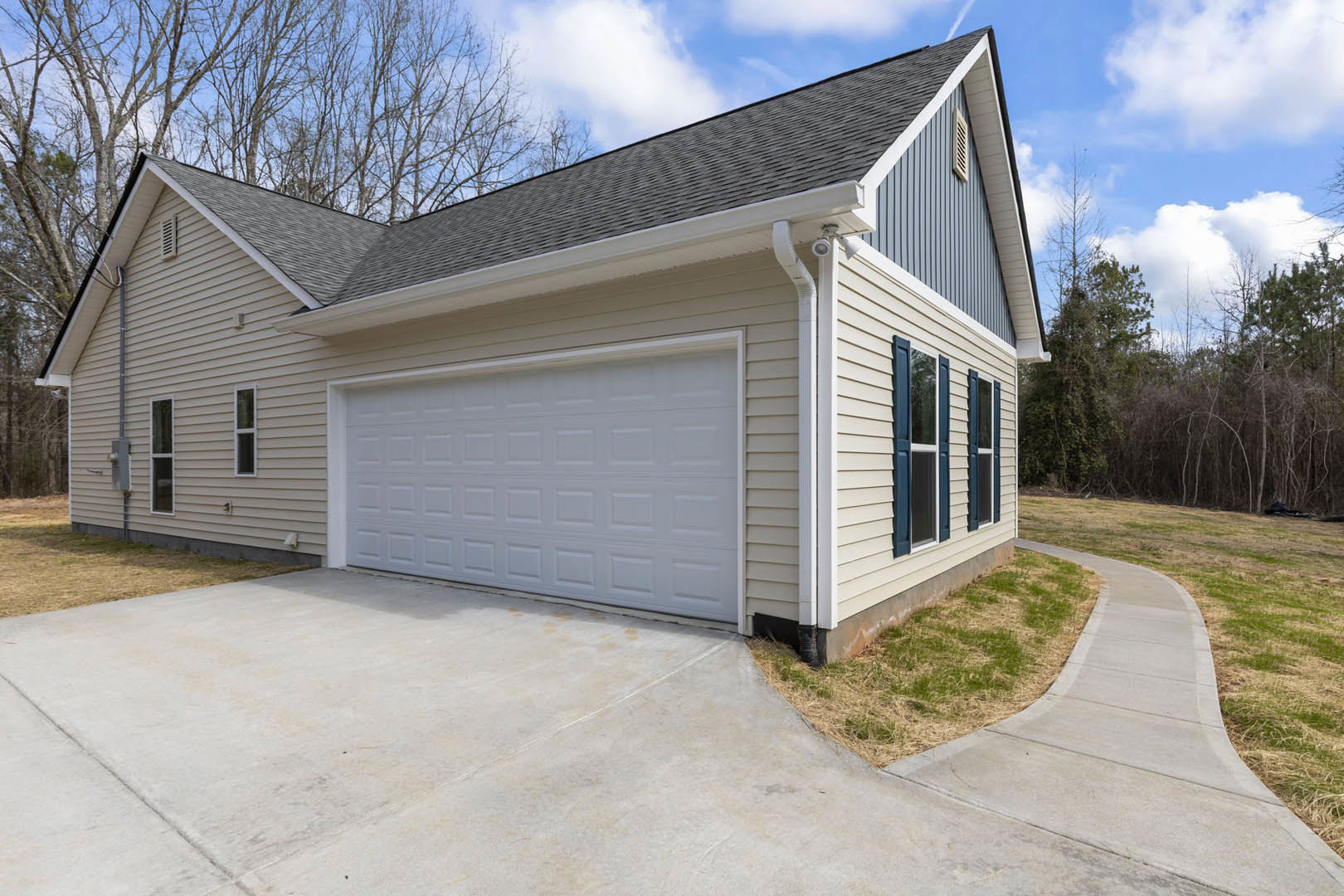 Two-story house with gray siding, white trim, attached garage, concrete driveway, and large windows; mature trees in background under partly cloudy sky