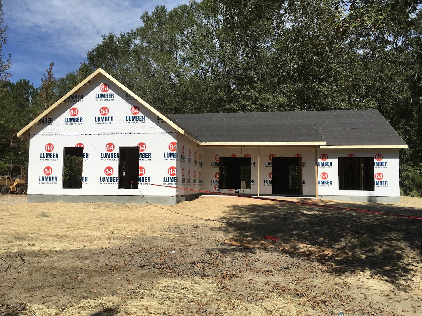 Wood-framed house under construction on dirt lot, surrounded by trees, with construction equipment and red safety tape visible