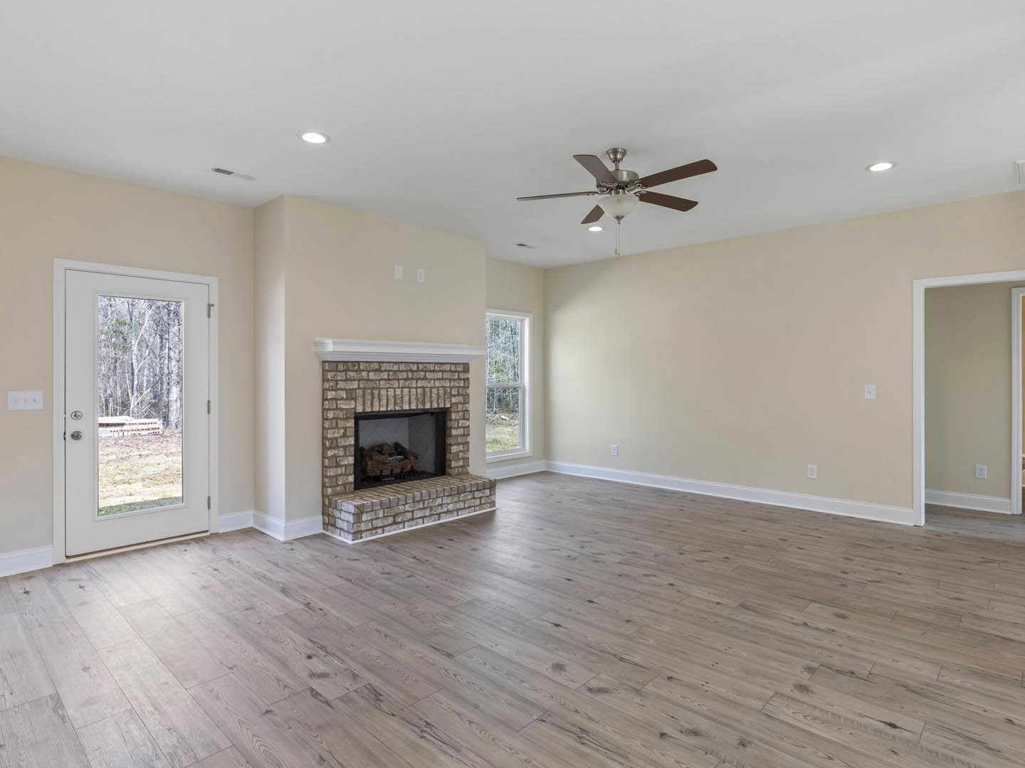 Living room with hardwood flooring, brick fireplace, ceiling fan with light fixture, white door frame, and door opening to forest view