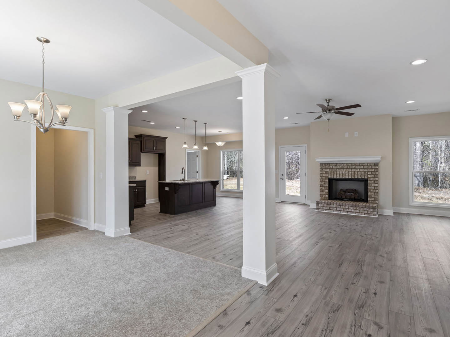 Open living room with laminate wood flooring, white framed fireplace, kitchen island with sink, white door with window, shelving holding a toy car, large window showing trees