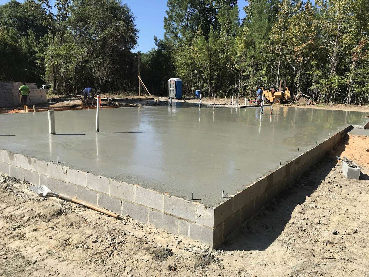 Concrete slab foundation with metal poles, portable toilet nearby, person in green shirt, surrounded by trees and dirt ground