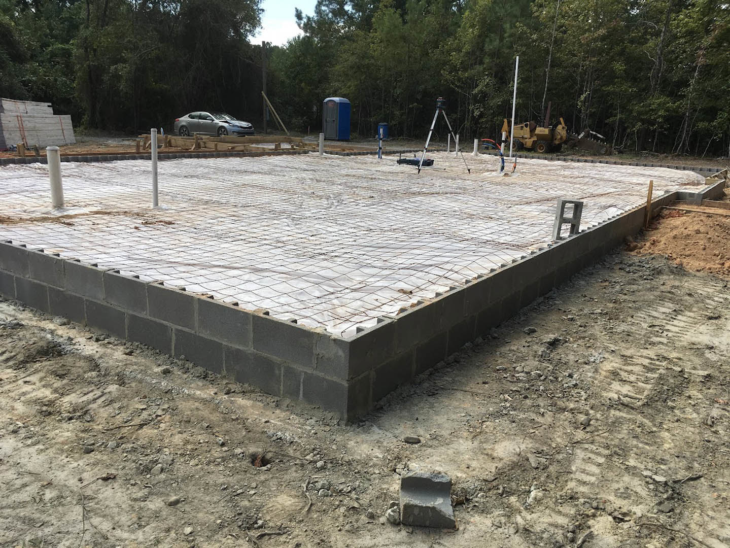 Concrete foundation with stacked cement blocks, grid pattern on ground, large yellow tractor with bucket, blue and white portable toilet, white wall with red ropes, car parked