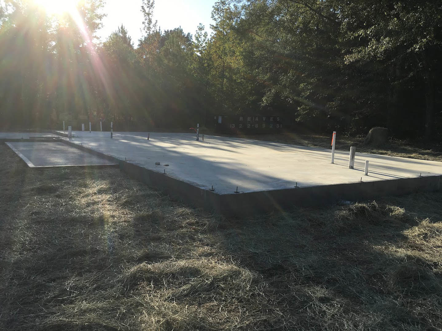 Concrete slab foundation with metal poles, surrounded by grass and trees in morning sunlight