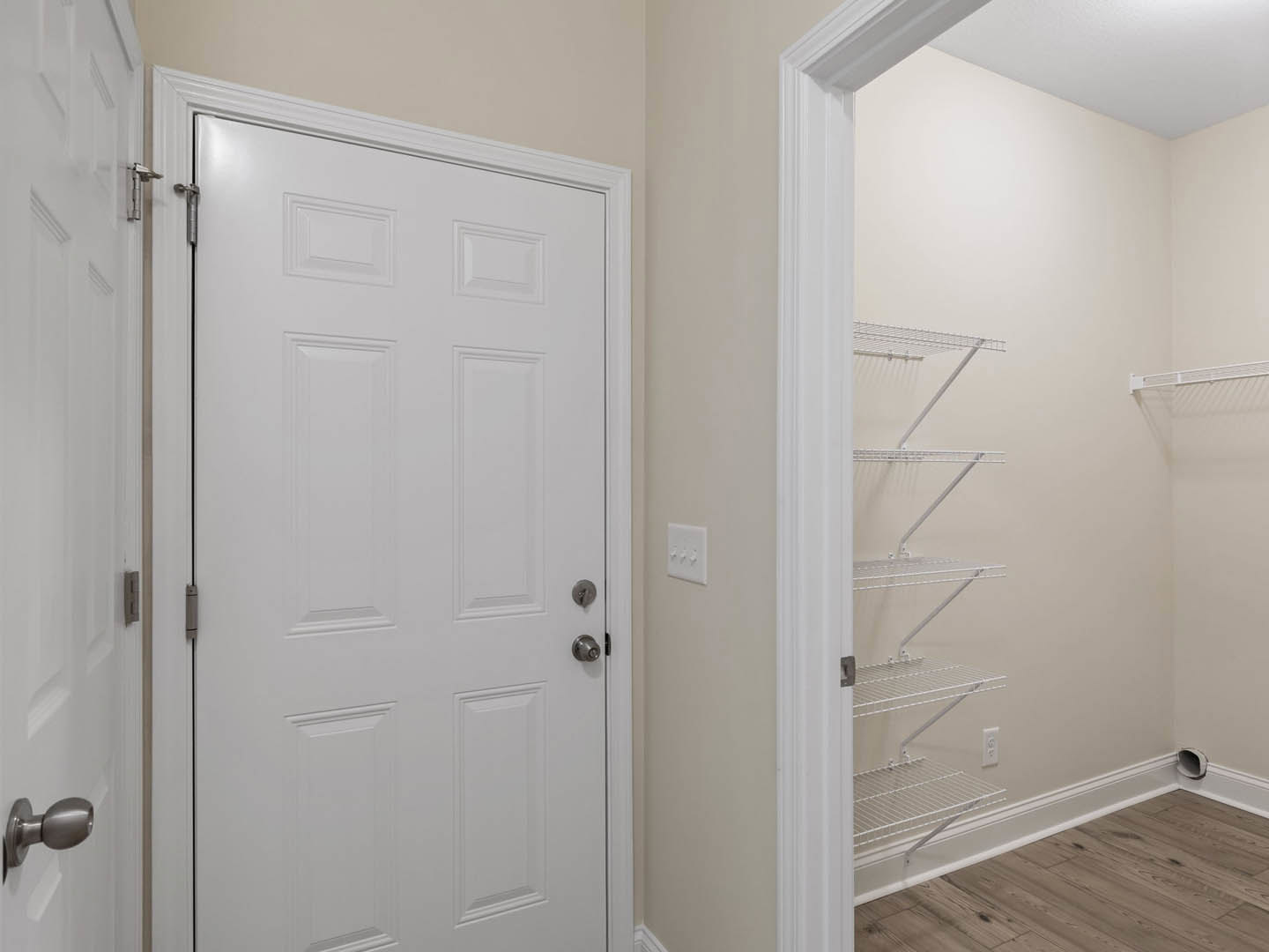 White paneled door with silver knobs and black trim in a hallway featuring wood flooring, white walls, a three-switch light plate, and a built-in white shelf.