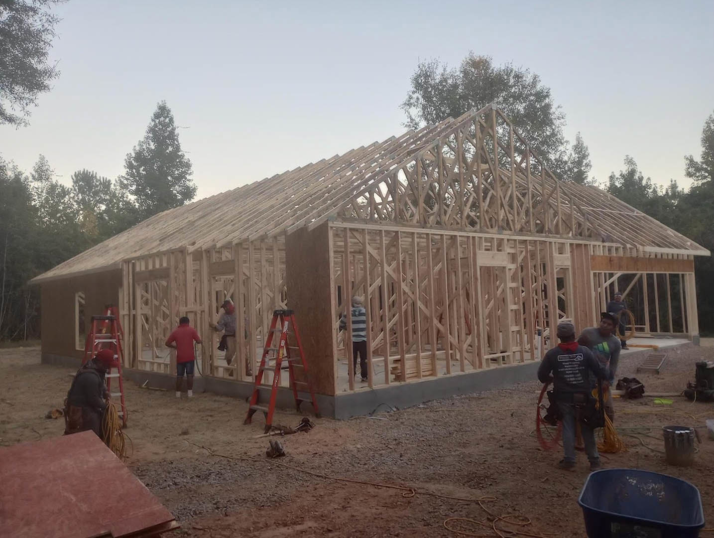 Construction site with workers assembling a modern home, exposed framing, black metal container, red construction materials, trees and sky in background
