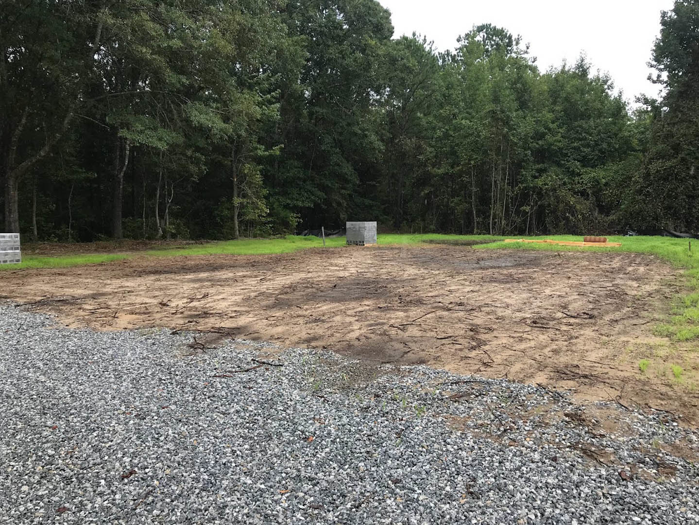 Dirt lot with scattered gravel piles, mature trees with green leaves in the background, open sky above