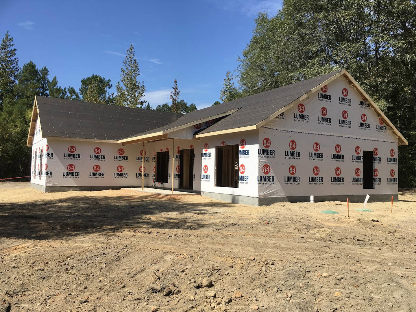 Wood-framed house under construction on a dirt lot, surrounded by leafy trees and cloudy sky in the background