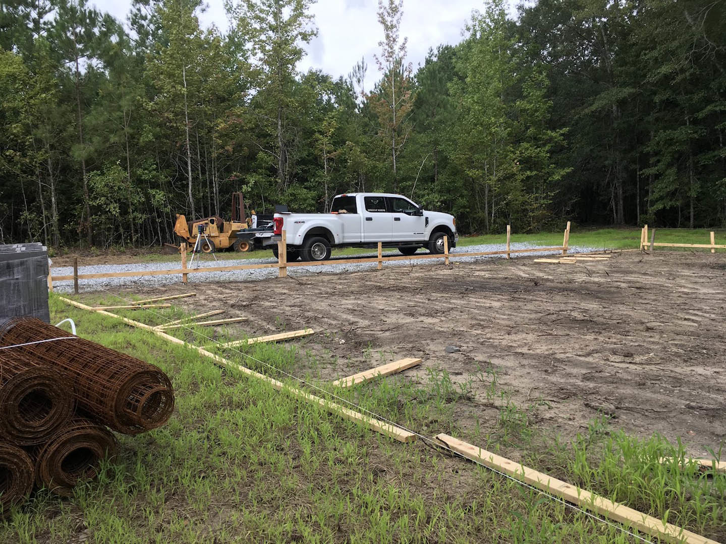 White pickup truck parked on dirt near metal rods and construction equipment, fenced construction site bordered by trees and grass, dog visible in truck window, man standing beside