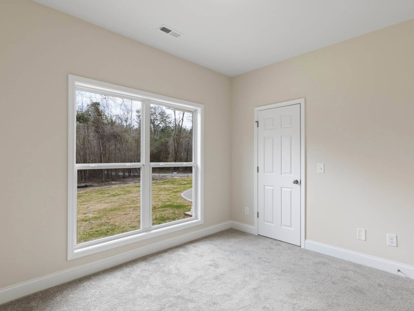 White carpeted room with a white door featuring a silver knob, large window showing grass and trees outside, and ceiling vent.