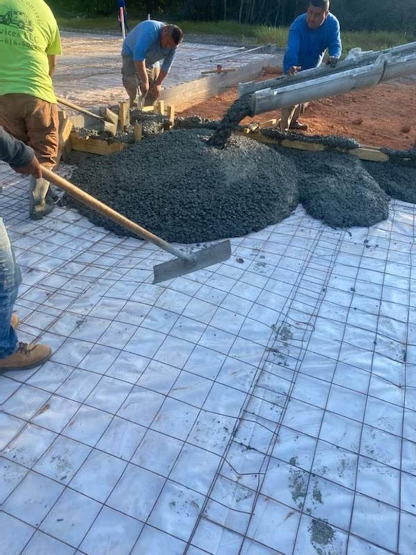 Men pouring concrete foundation outdoors, one wearing a green shirt and another in blue, with a shovel resting on the grey surface and visible construction materials.