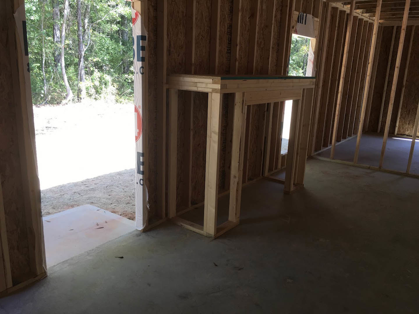 Exposed wooden framing and beams in a partially constructed room with a concrete floor, large windows, and views of surrounding trees