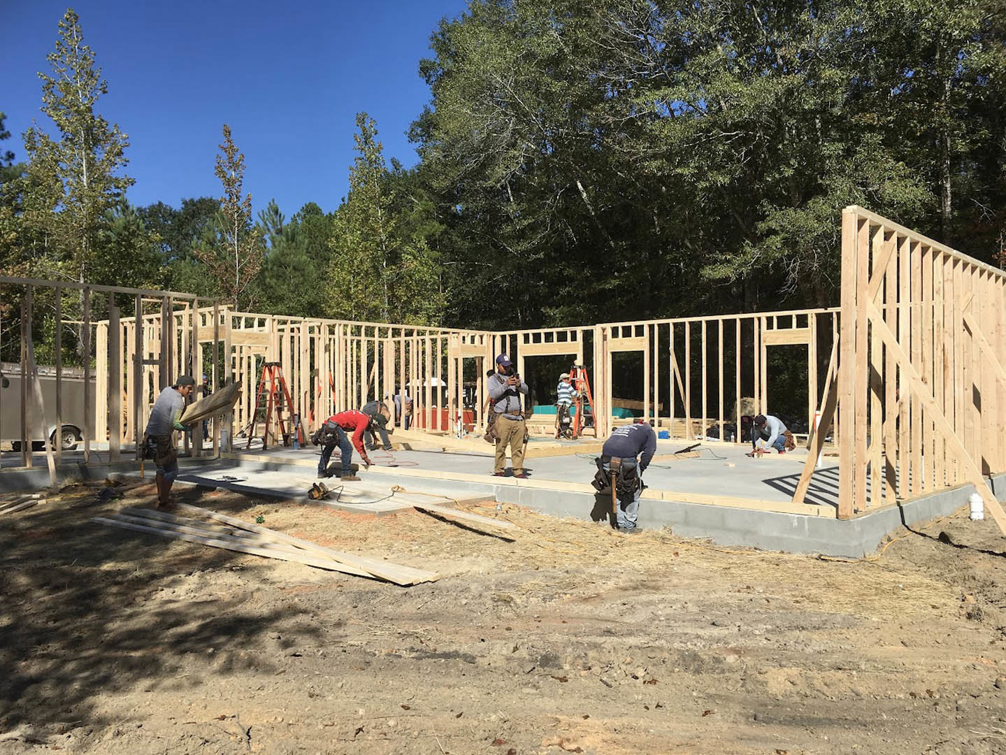 Framed wooden exterior wall with a cross brace, pile of lumber on dirt ground, three men working—one with a tool belt, one bending near a curb, one holding a camera—trees and blue