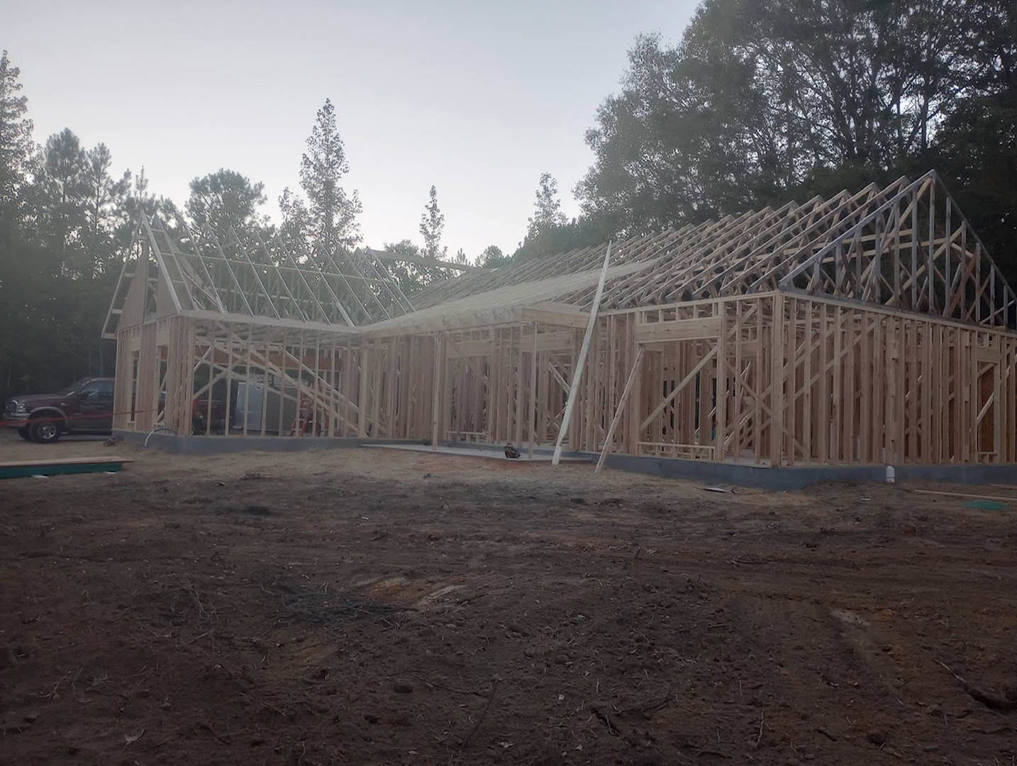 Wood-framed custom home under construction on a dirt lot, surrounded by leafy trees and a wooden fence, with exposed beams and a car parked nearby.