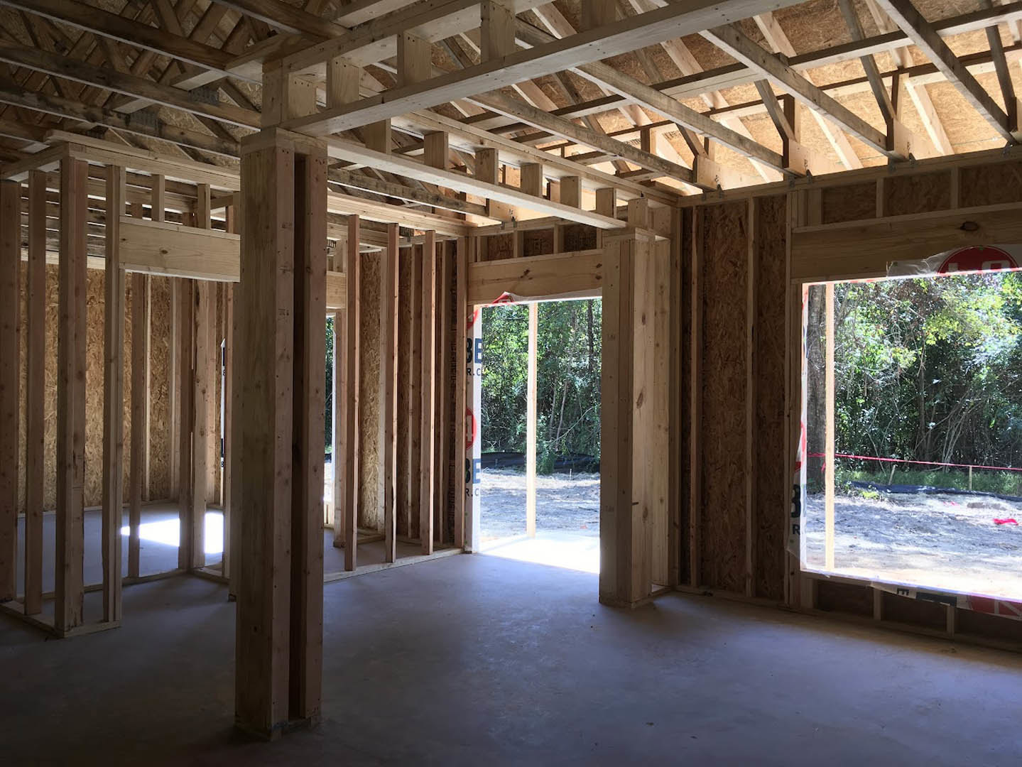 Open room with exposed wood ceiling beams, large window framing leafy trees, concrete floor illuminated by natural daylight, and a simple door.