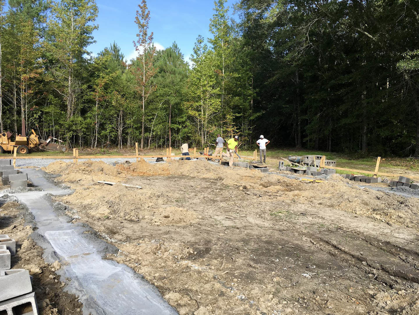Group of people standing on bare soil at a residential construction site near a dirt road, yellow tractor and trees in background