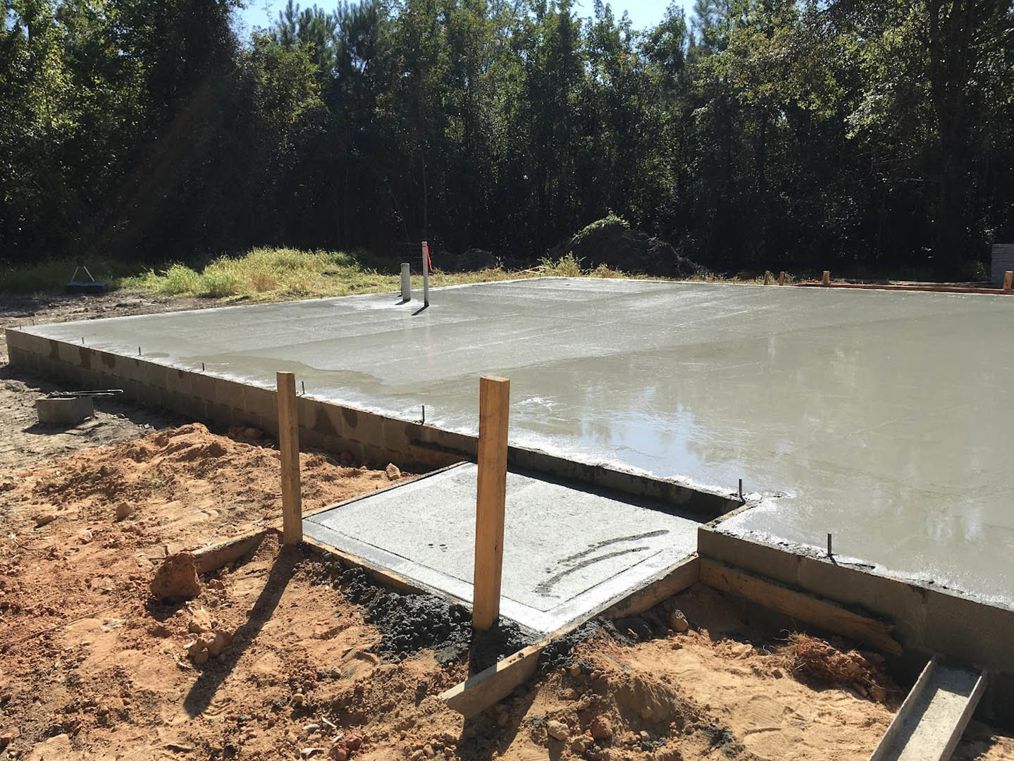 Freshly poured concrete slab foundation with wooden posts, surrounded by trees and construction materials on bare ground