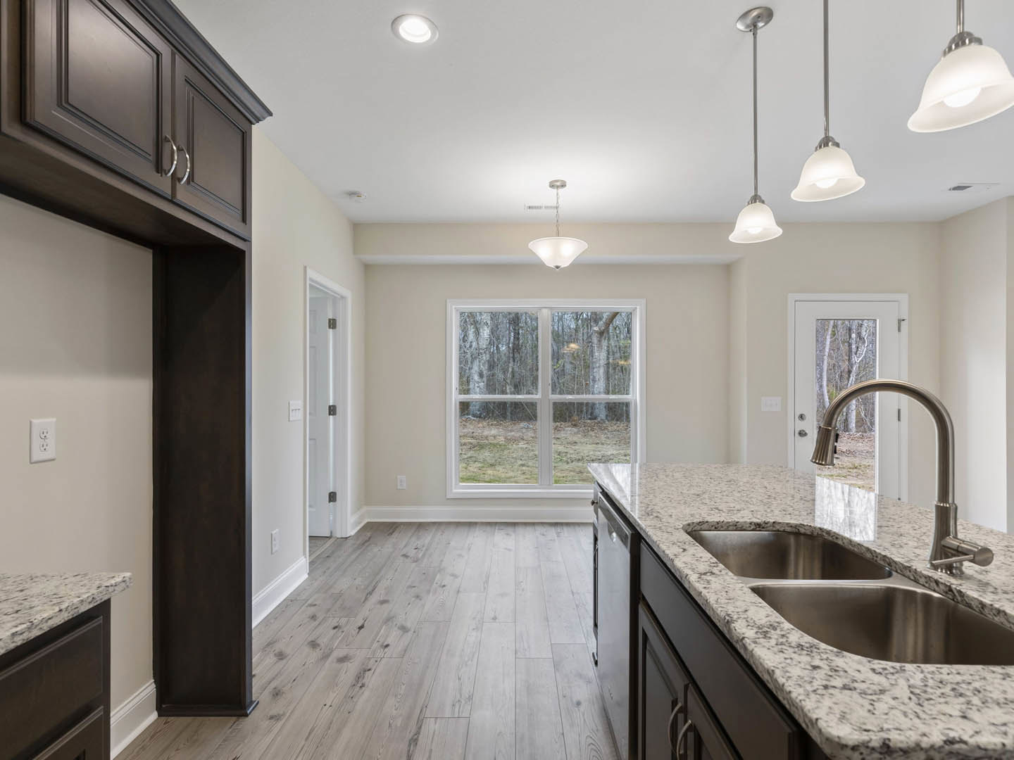 Kitchen with wood flooring, double stainless steel sink set in a stone countertop, white cabinetry, ceiling light fixture, window overlooking trees, and chrome faucet.