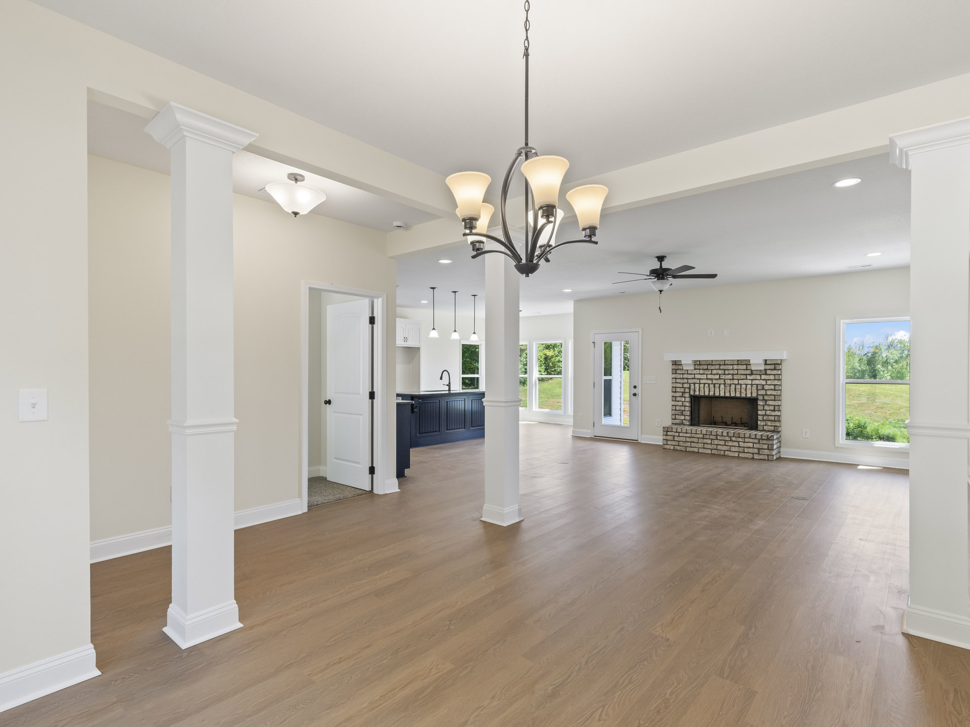 Spacious living room featuring wood flooring, white pillars, brick fireplace, chandelier with light fixture, large window overlooking trees, and glass-paneled white door