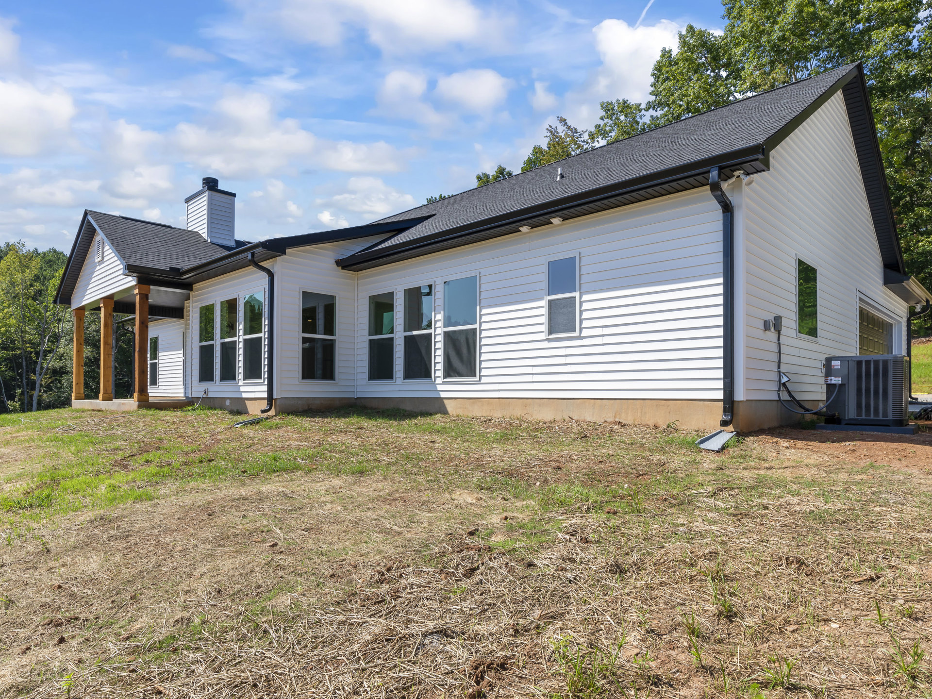 White house with black roof, large windows with white frames, covered porch, green lawn in foreground, mature trees in background, cloudy sky overhead
