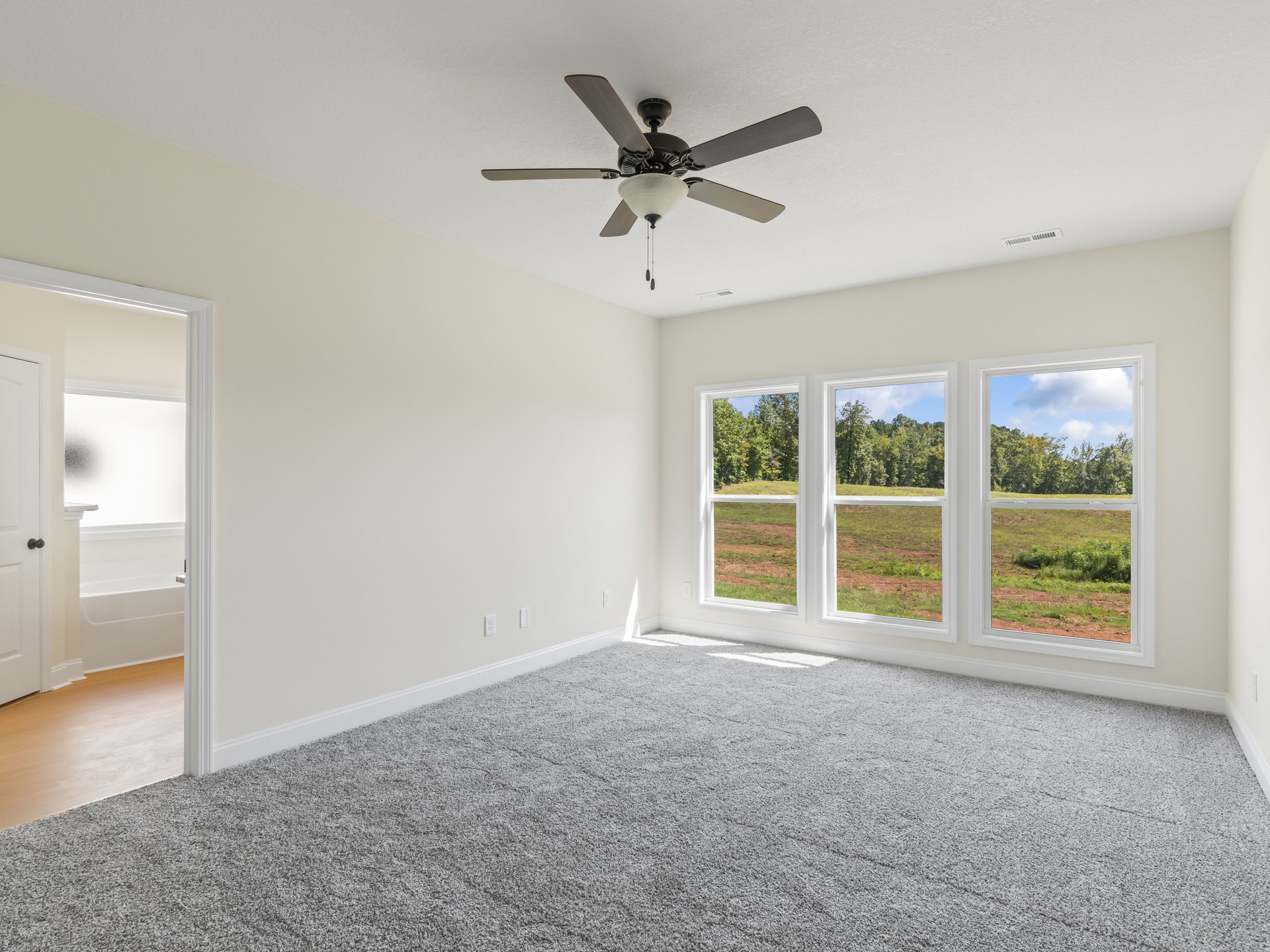 Ceiling fan with light fixture above carpeted floor, large windows with tree views, neutral walls, and white trim