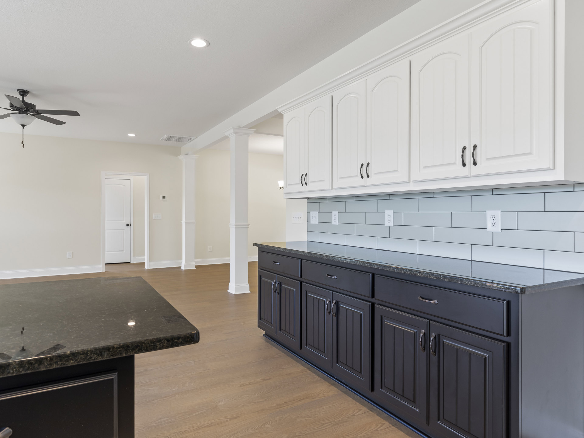 White kitchen cabinets with black handles, black countertops, white tile backsplash, ceiling fan with light, white door with black knob, stainless steel sink, and black kitchen