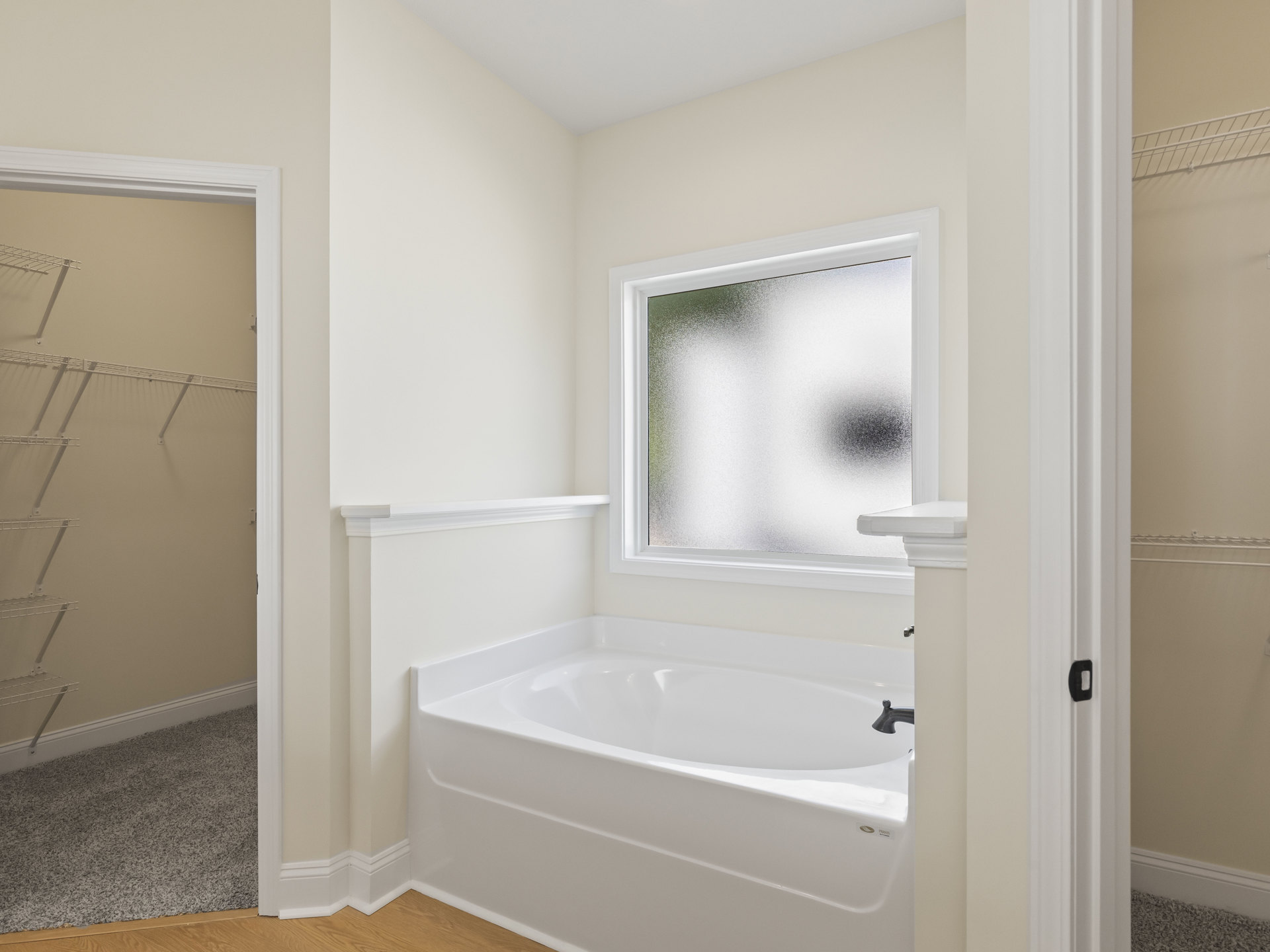 Freestanding white bathtub on wood flooring beside frosted window, white wall with metal shelves, neutral carpet, and chrome tap fixtures