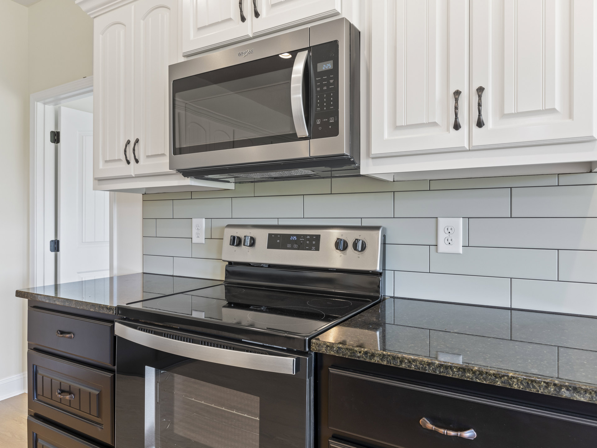 White kitchen cabinets with black countertops, glass-top stove, built-in microwave, brushed metal handles, electrical outlet, and wall clock