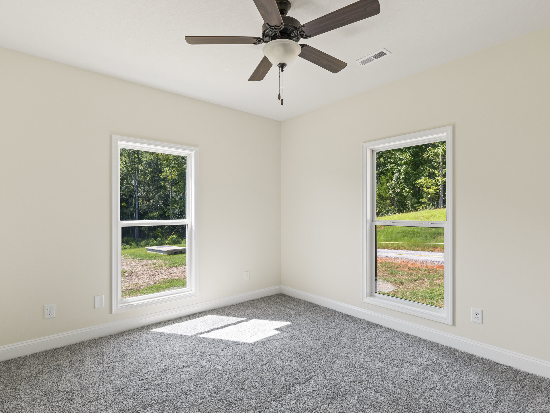 Ceiling fan with light fixture above white and grey carpeted floor, large windows showing grassy yard and trees, white plaster walls