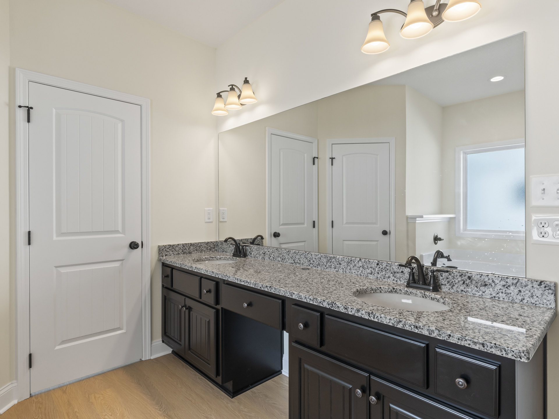 Bathroom featuring double sinks set in a granite countertop, large wall mirror, white cabinetry with black handles, frosted glass window, and a three-light fixture above the