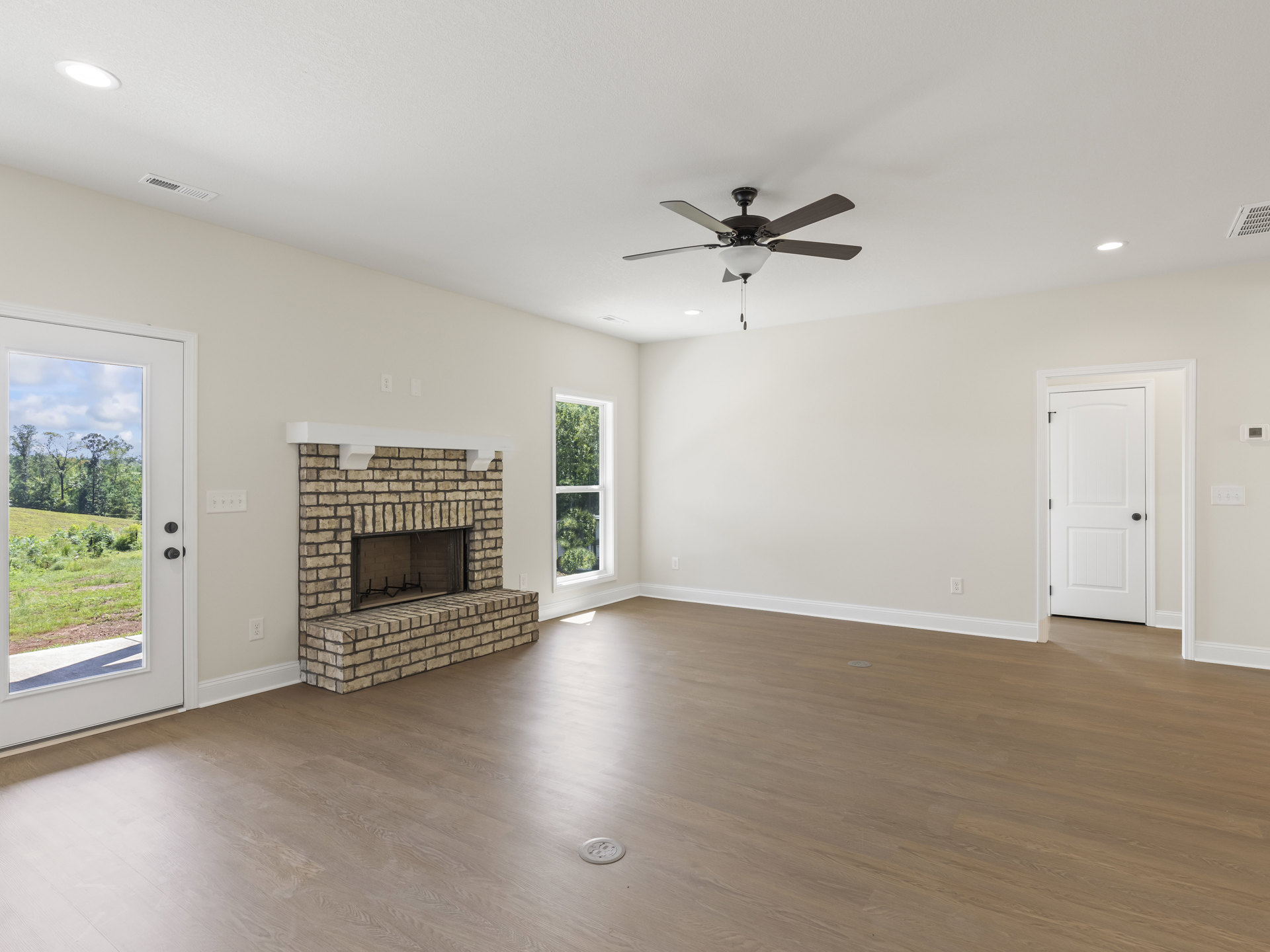 Living room with wood flooring, white-framed fireplace, ceiling fan with light fixture, white door with black knob, circular object on floor, door opening to view of field and