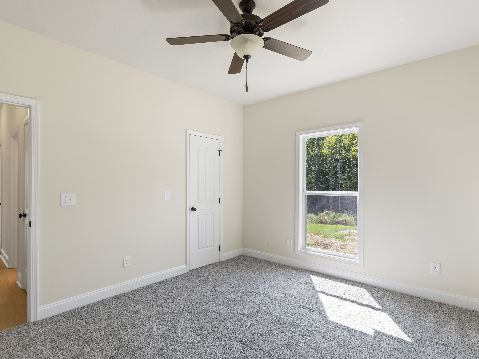 Carpeted room with white walls, ceiling fan with light fixture, window showing trees outside, white door with black knob, light switch on wall