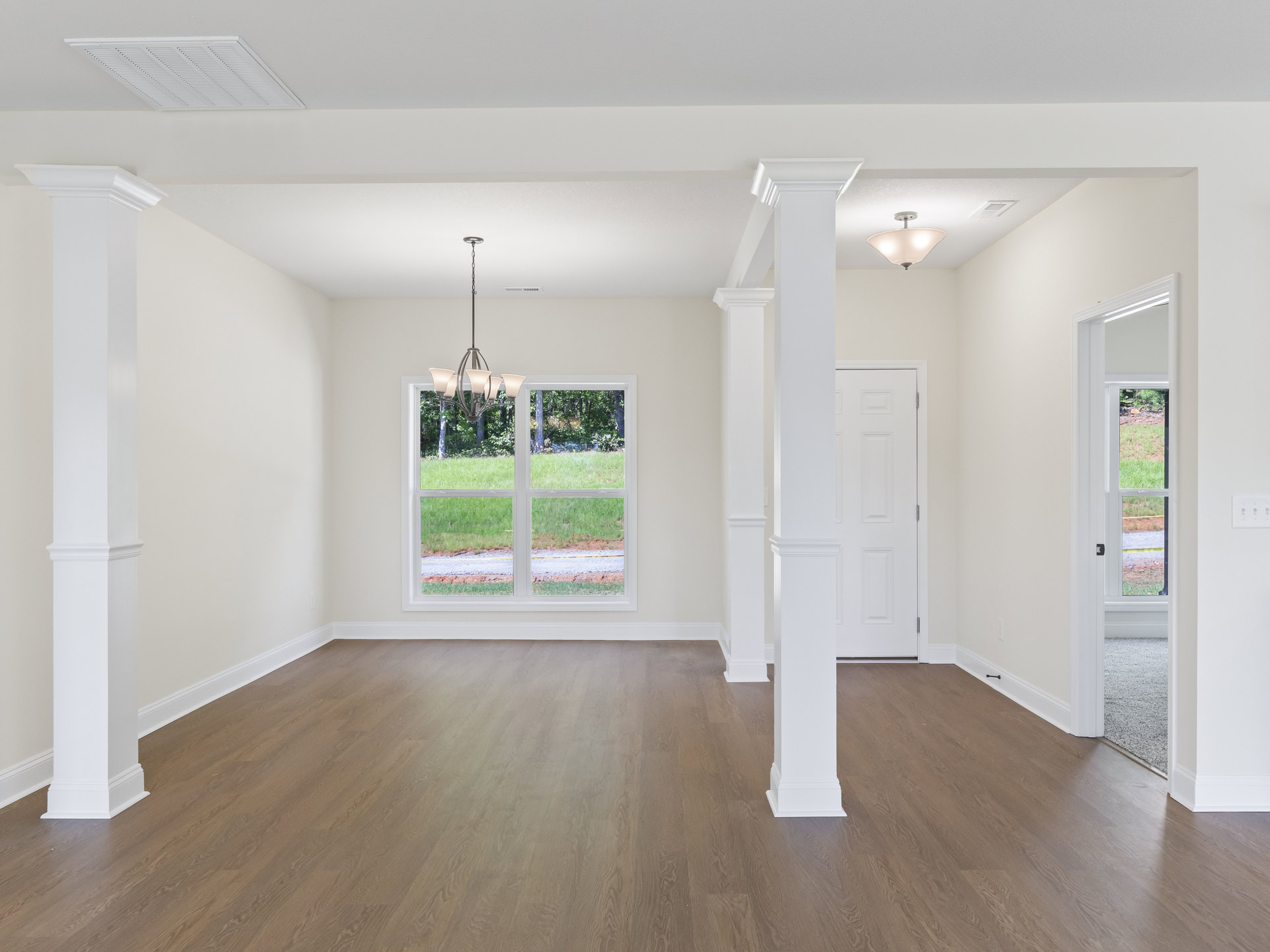 Wood flooring with white pillars, white walls, a white door with metal frame, window showing grass outside, ceiling light fixture