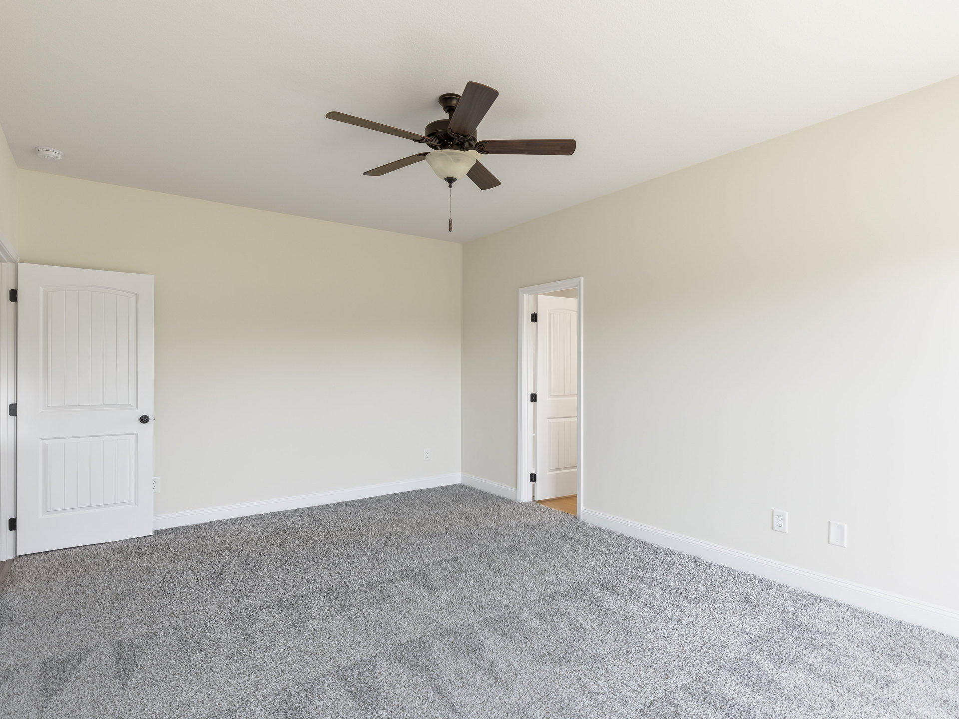 Carpeted bedroom with white walls, ceiling fan with light fixture, white door featuring black knob and visible hinge
