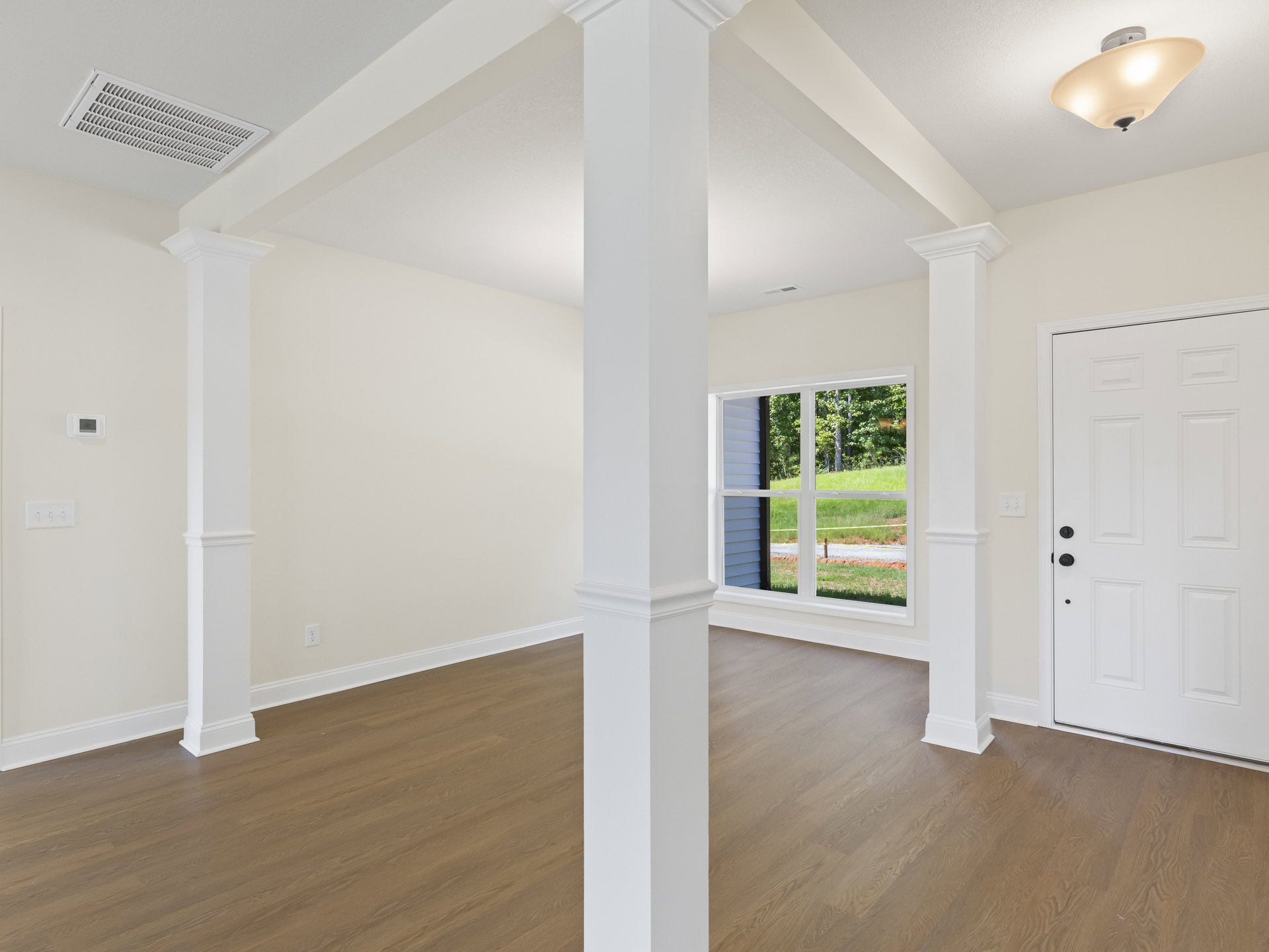 White pillar stands in a room with laminate flooring, white walls, a ceiling vent, modern light fixture, white door with black knobs, and window overlooking a grassy field.