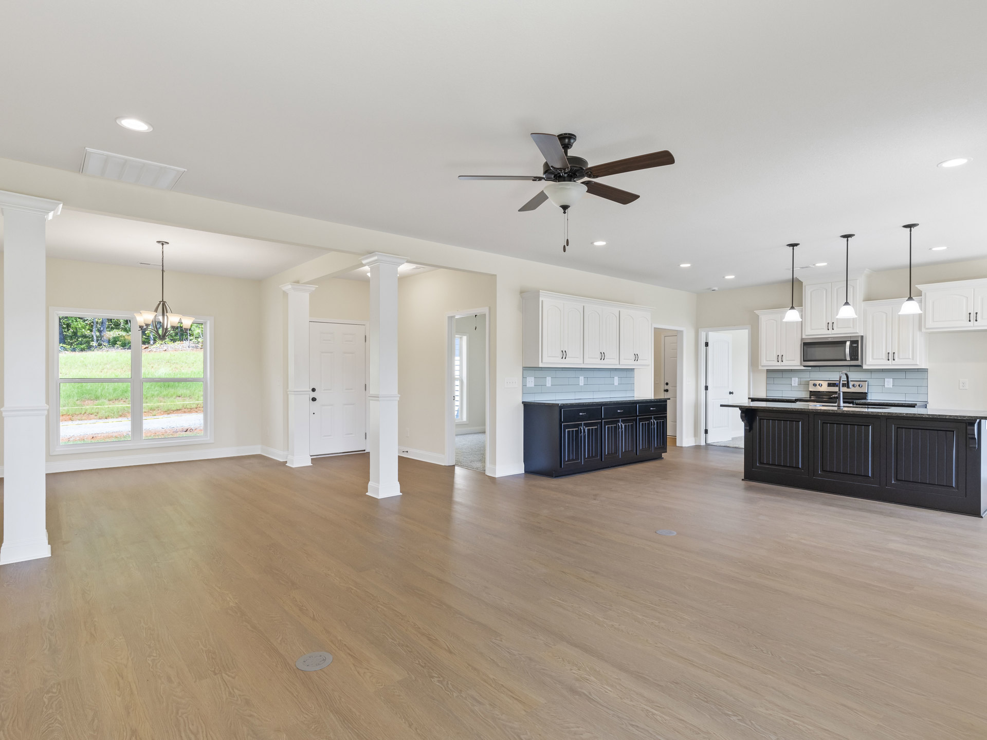 Spacious open floor plan featuring wood flooring, black kitchen cabinets and countertops, ceiling fan with light fixture, large window overlooking grassy yard