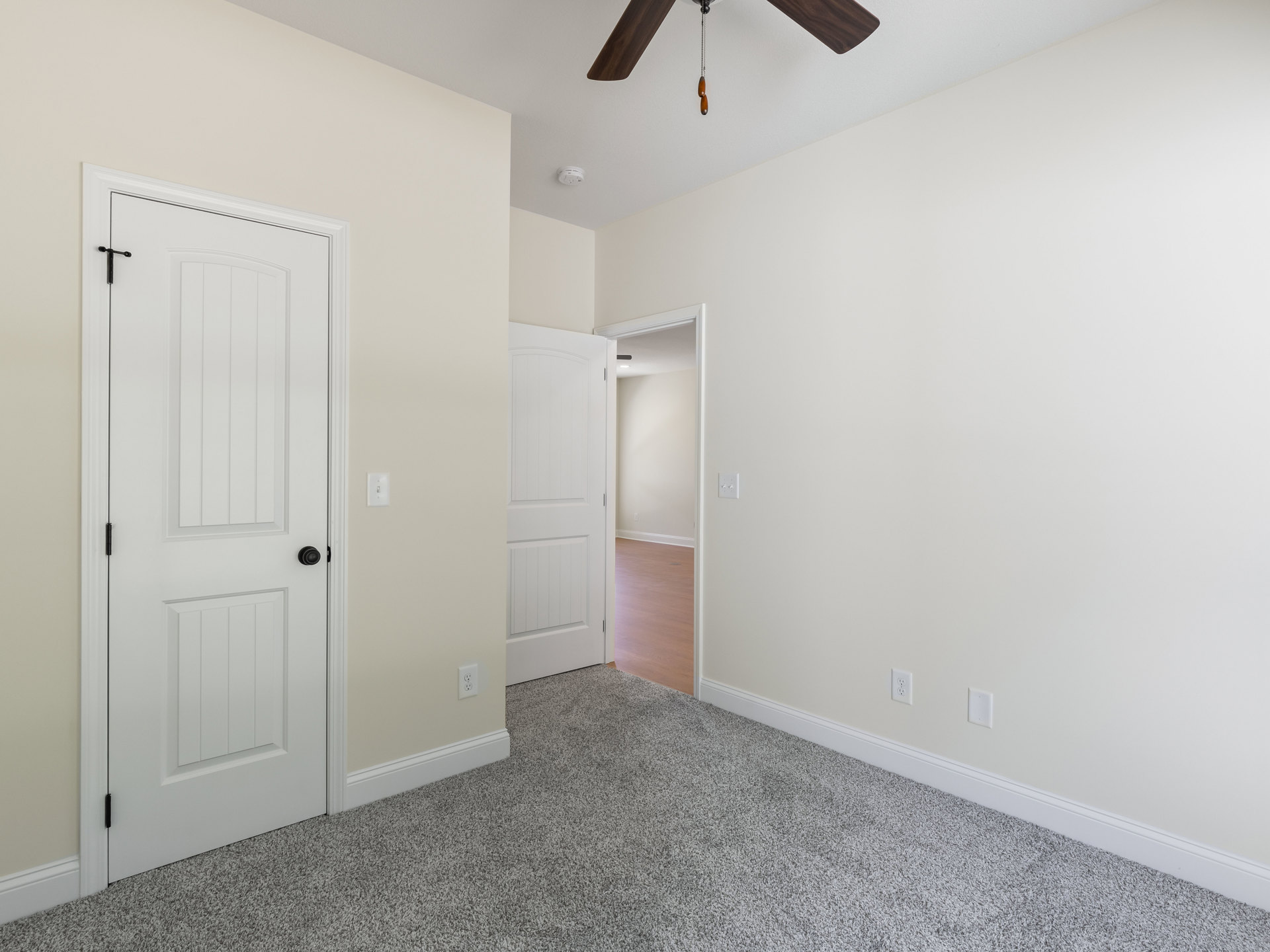 Ceiling fan mounted on white plaster ceiling, white door with black knob, wood flooring transitioning to carpet, white walls.