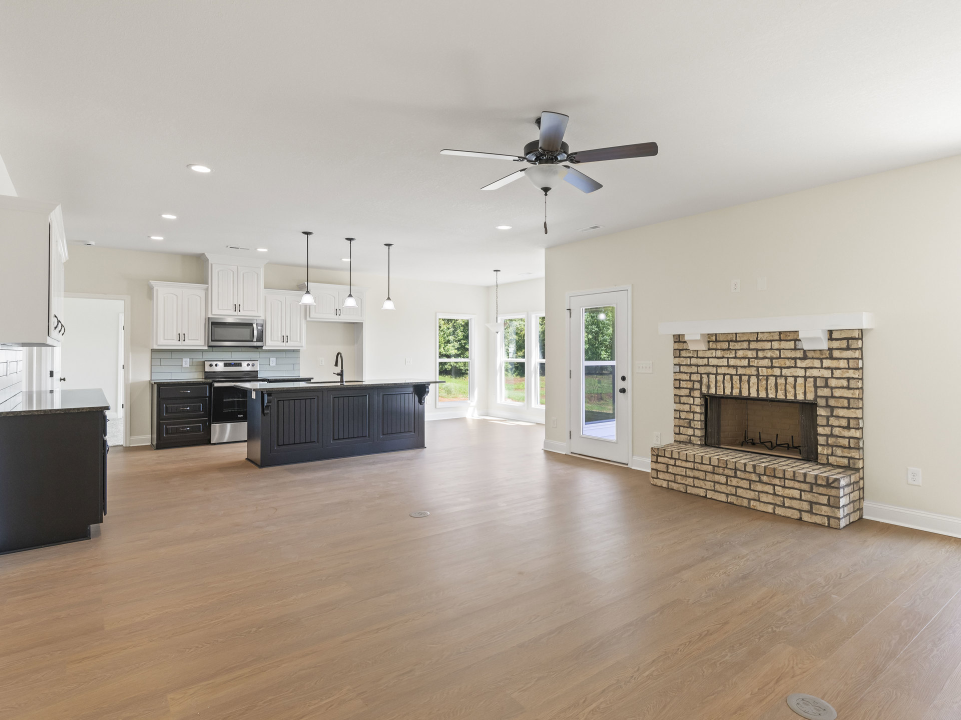 Open-concept living room with stone fireplace, ceiling fan with light, adjoining kitchen featuring black countertops, cabinetry, microwave, and screened door.