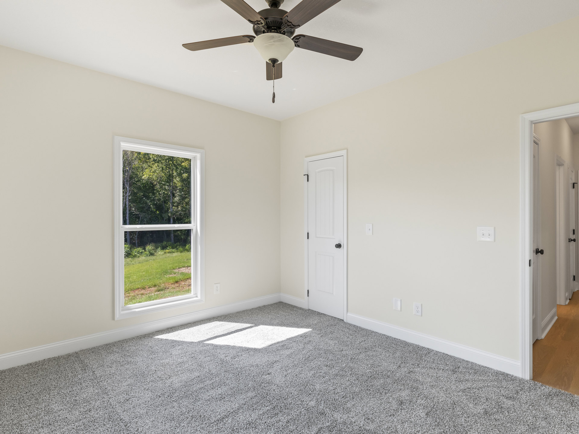 Carpeted room with white walls, grey and white flooring, ceiling fan with light fixture, large window overlooking forest, white door with black knob