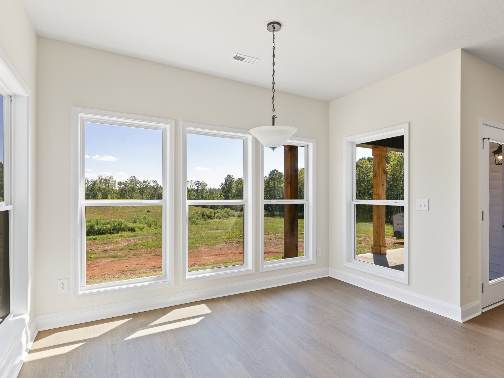 Sunlit room with large windows overlooking a grassy field and trees, featuring wood flooring and white walls