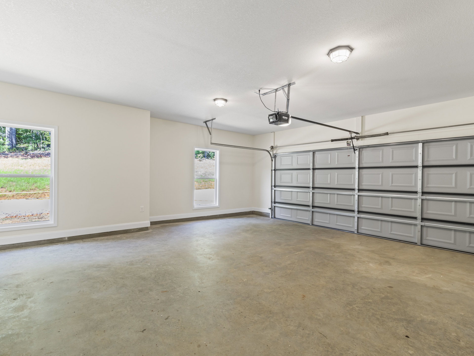 Spacious garage with white plaster walls, concrete floor, large window overlooking grass and train tracks, ceiling light fixture, and metal-framed garage door