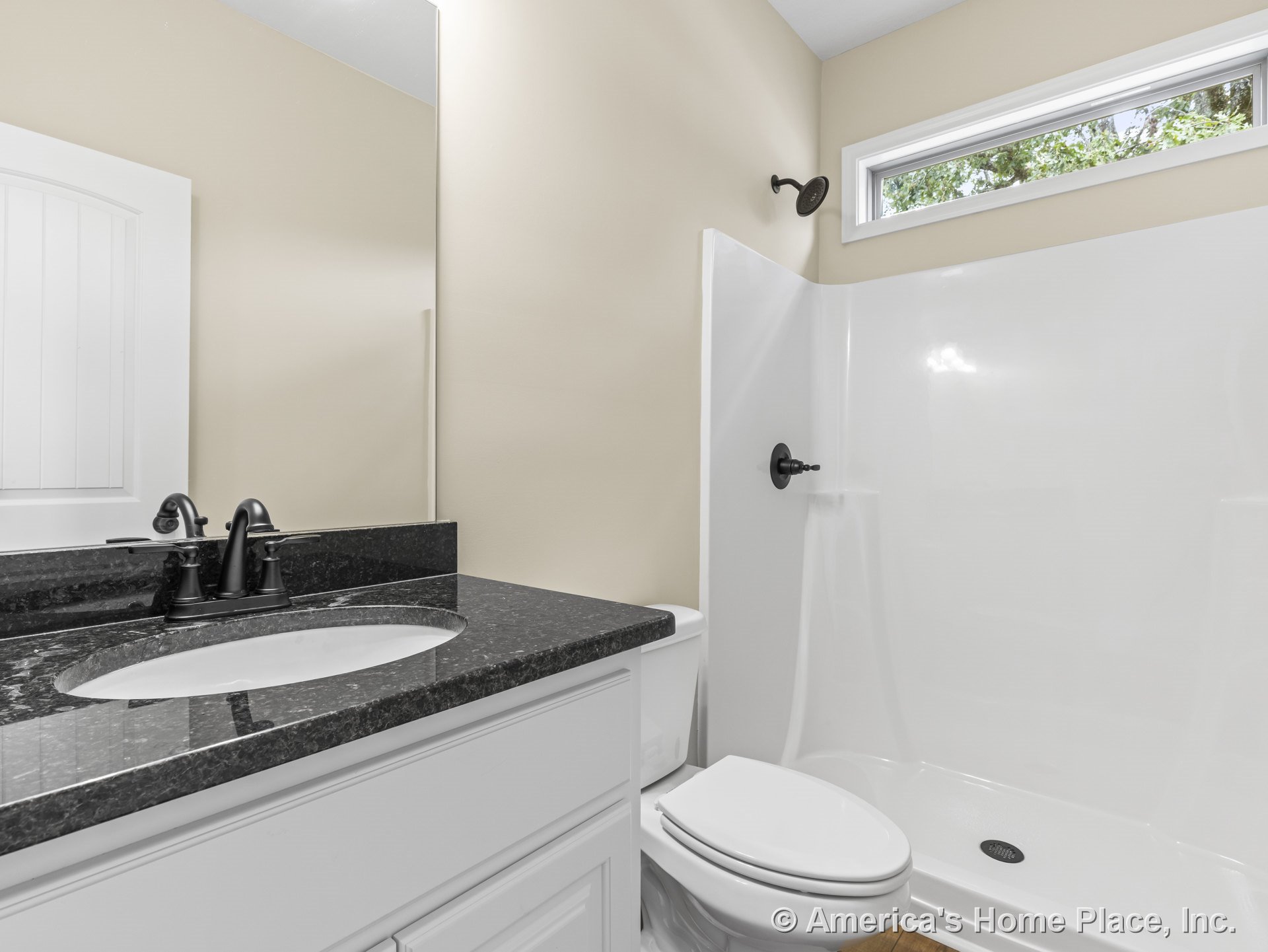 Walk-in shower with black showerhead and faucet, single white vanity with black granite countertop, large wall mirror, rectangular window above shower, light beige painted walls.