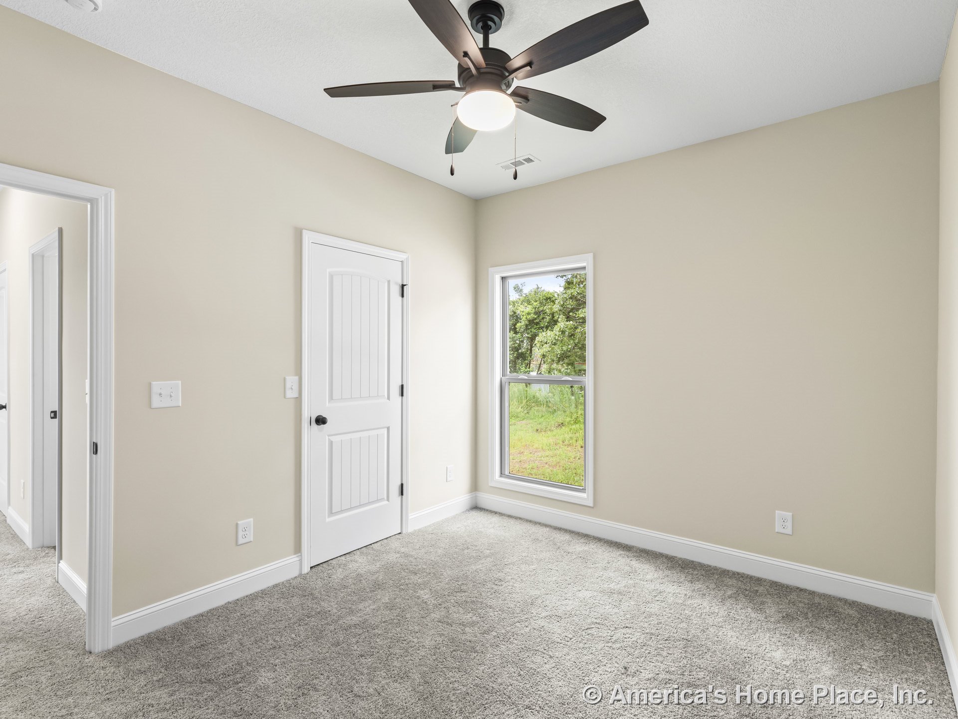 Bedroom with beige walls, carpet flooring, single window framed in white casing, white paneled door, white baseboards and trim, and ceiling fan with integrated light fixture.