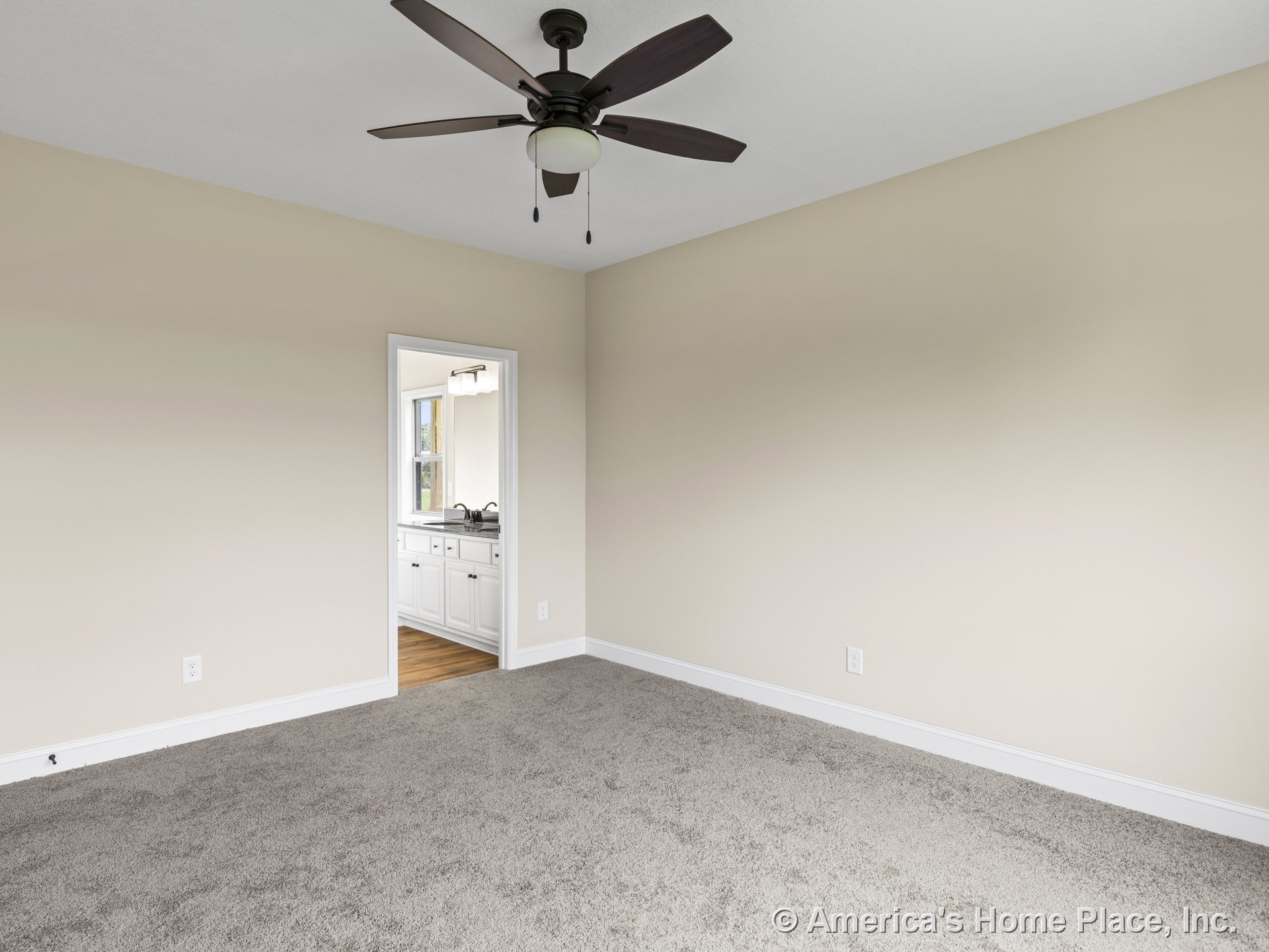 Carpeted bedroom with neutral walls, ceiling fan, white baseboard trim, and open doorway revealing bathroom with white cabinetry and window.